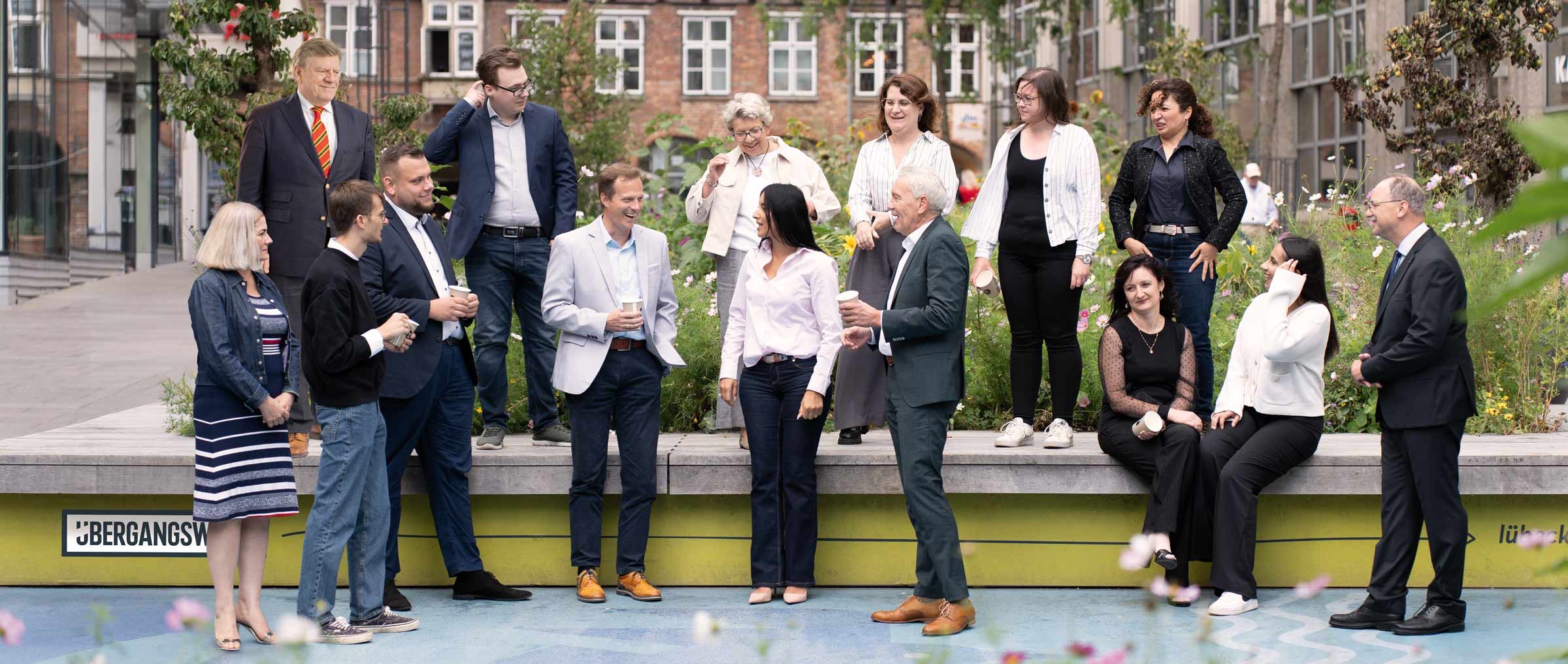 Das Bild zeigt eine Gruppe von ARGON-Mitarbeitern, die locker und freundlich im Freien zusammenstehen und sitzen, teils im Gespräch miteinander. Der Hintergrund besteht aus modernen und älteren Gebäuden sowie Grünflächen mit Blumen. Die Personen sind überwiegend geschäftlich gekleidet, wobei ein Mann im Anzug auf einem Podest auffällig wie eine lebensgroße Pappfigur wirkt. Die Atmosphäre wirkt entspannt und gemeinschaftlich.