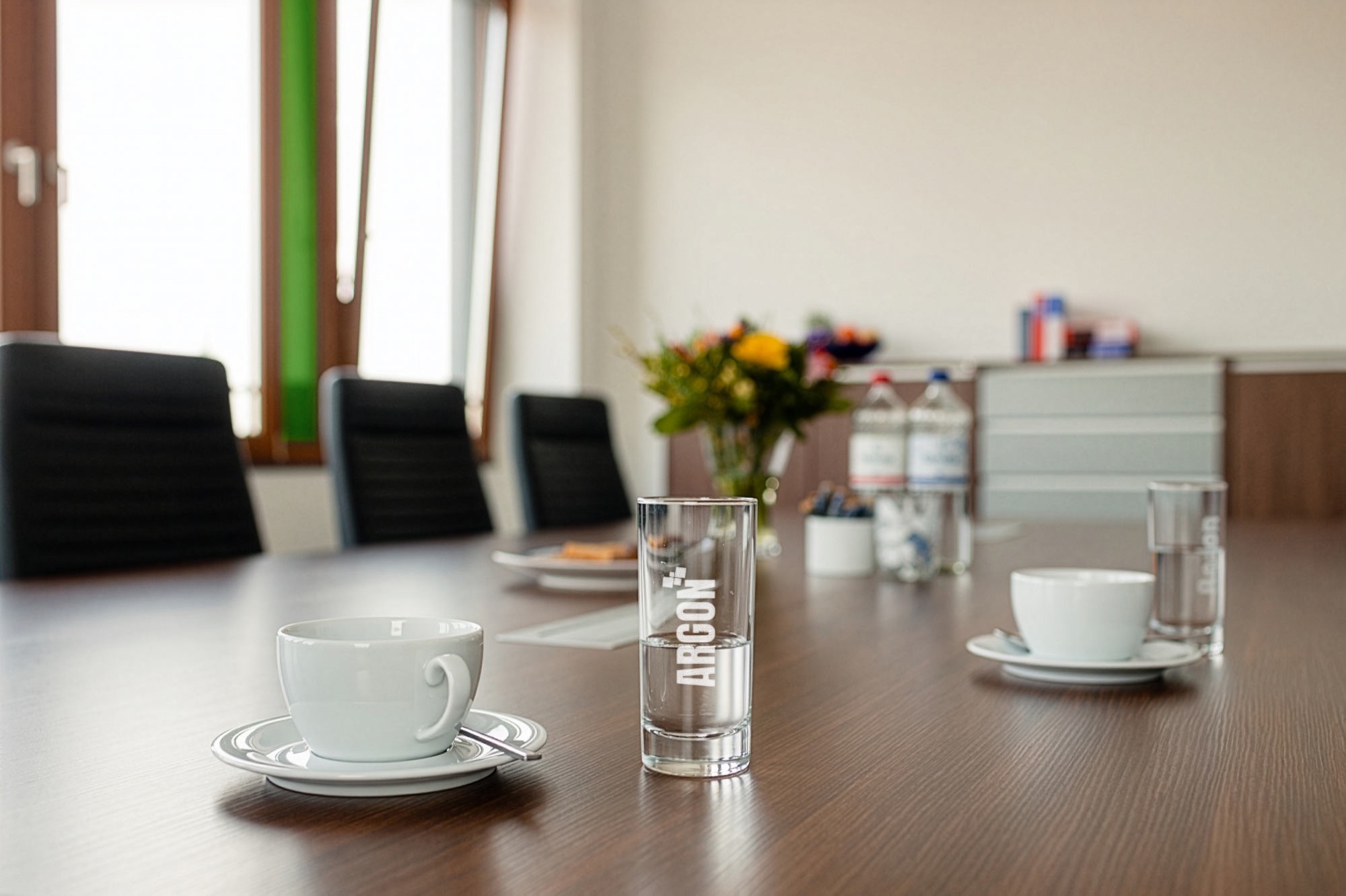 Das Bild zeigt einen modernen Konferenzraum mit einem langen Holztisch, auf dem mehrere weiße Kaffeetassen mit Untertassen sowie Wassergläser stehen. Im Hintergrund sind schwarze Konferenzstühle, große Fenster mit Tageslicht und ein Sideboard mit Blumenstrauß, Wasserflaschen und Büroartikeln zu sehen. Die Atmosphäre wirkt einladend und professionell.