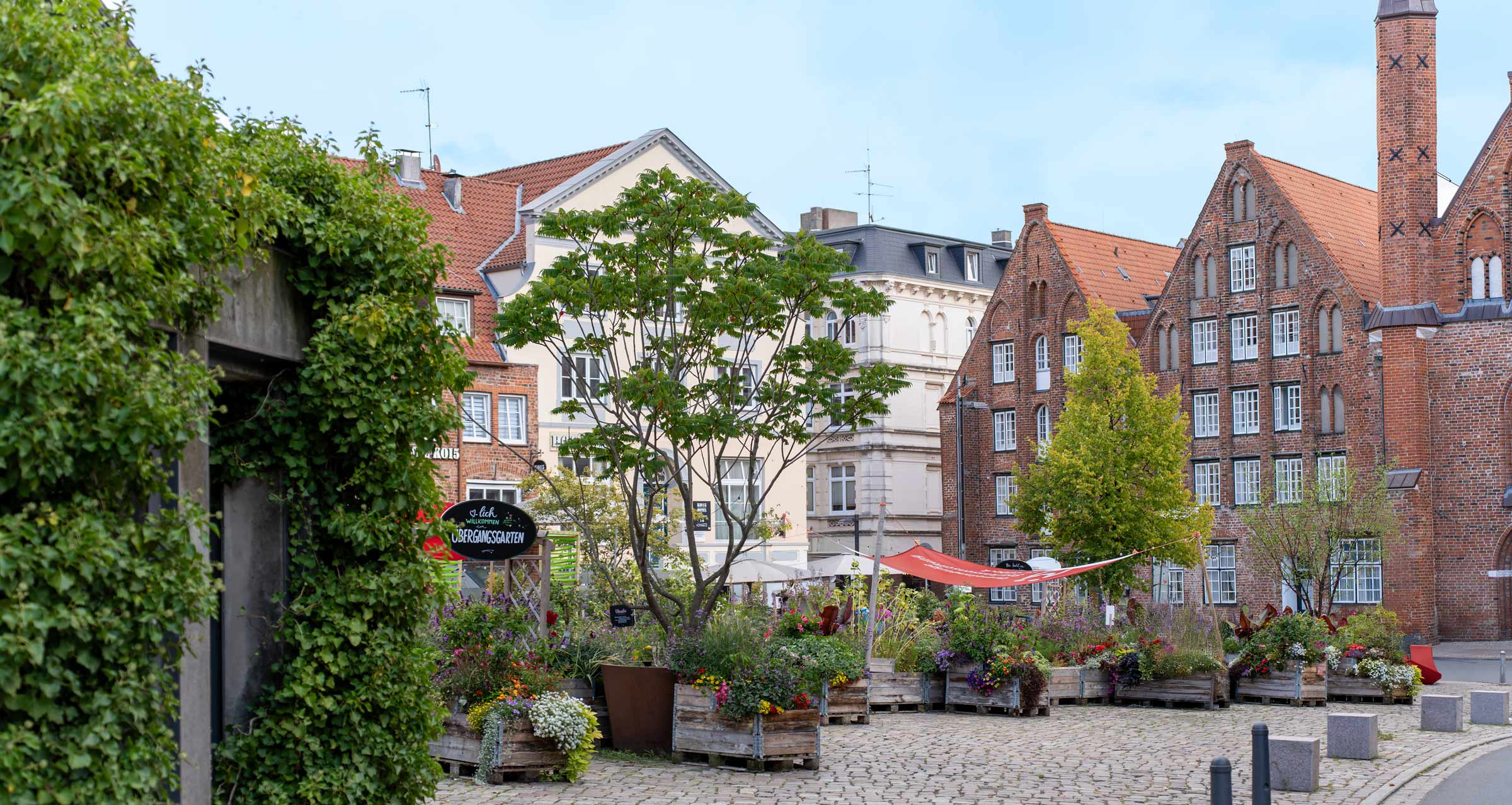 Bunter Stadtgarten mit Hochbeeten und Blumen auf einem gepflasterten Platz, umgeben von historischen Backsteinhäusern mit Giebeln und roten Ziegeldächern.